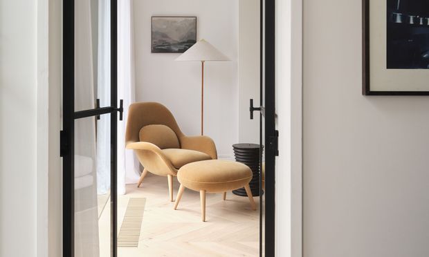 A refined sitting area viewed through open black-framed glazed doors. The room features a sculptural tan lounge chair with matching ottoman, a slim floor lamp, framed artwork, a dark side table, and pale herringbone timber flooring. The composition is bright and calm, with a restrained mid-century-inspired interior.