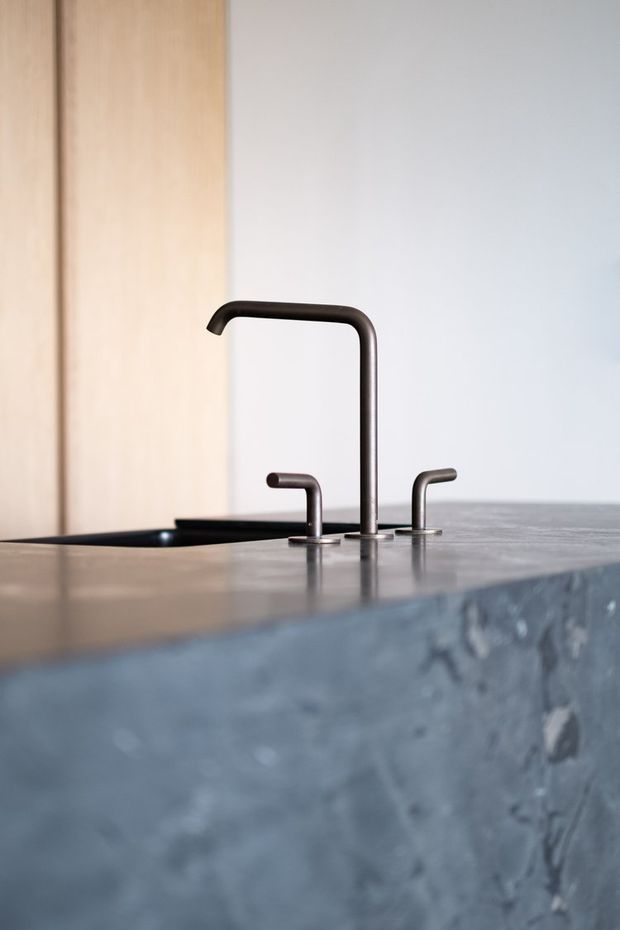 Close-up of a minimalist kitchen island with a dark metallic gooseneck mixer tap and two smaller matching fixtures set into a smooth grey benchtop. A black undermount sink is partially visible, with pale timber cabinetry or panelling in the background and a textured blue-grey island face in the foreground.