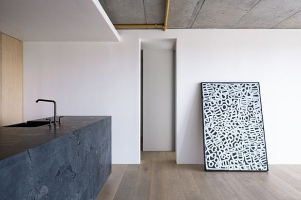 Minimal contemporary open-plan kitchen interior with a dark stone island and integrated sink, pale timber cabinetry, light timber flooring, white walls, and an exposed concrete ceiling with visible services. A narrow hallway or doorway is centered at the rear, while a large monochrome artwork leans against the wall.