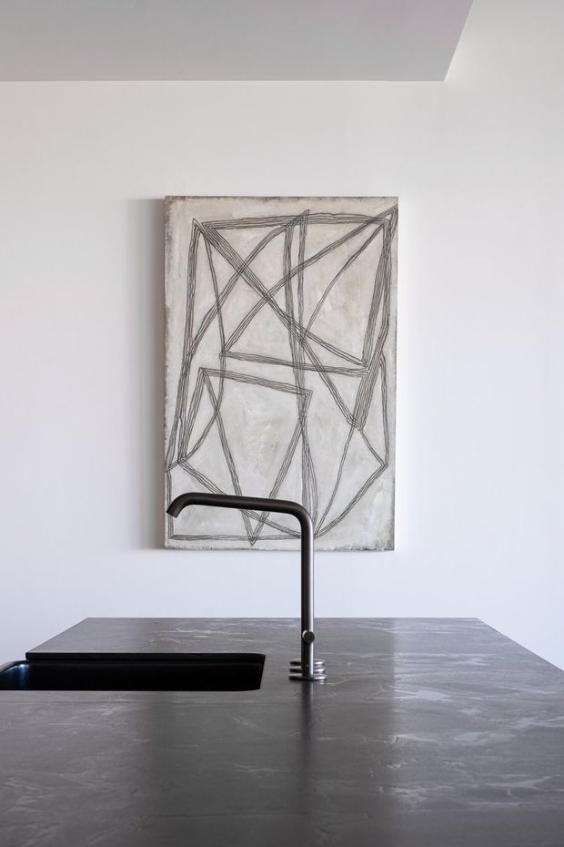 A minimalist kitchen view focused on a dark stone island benchtop with an integrated black sink and sculptural dark metal tap. A large abstract linear artwork hangs on the white wall behind, with a recessed ceiling detail visible above.