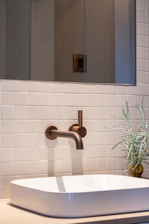 Close-up of a contemporary bathroom vanity with a white vessel basin, wall-mounted brushed bronze tapware, pale square tiled wall, and a large frameless mirror. A small decorative plant adds greenery beside the basin.