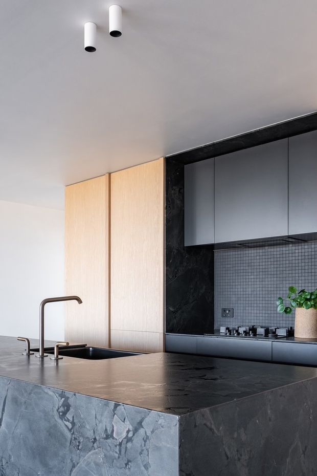 Contemporary kitchen featuring a large dark stone island with integrated sink and bronze-toned taps, matte dark cabinetry, a small square-tile backsplash, black stone wall panels, and tall light timber pantry cabinetry. Minimal cylindrical ceiling lights and a potted plant complete the refined, monochromatic scheme.