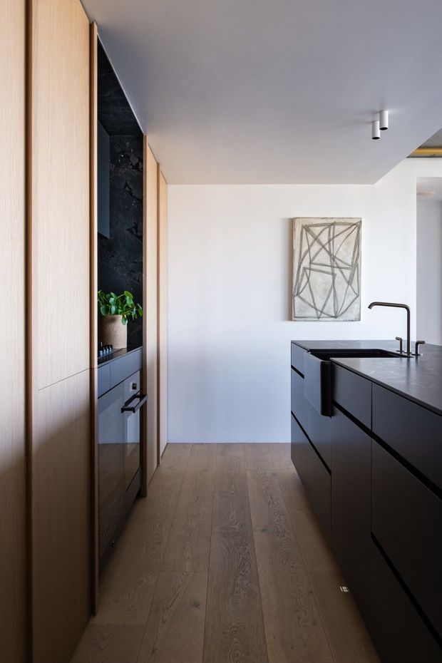 A refined contemporary kitchen with a narrow galley-style layout. Tall light timber cabinetry lines the left wall, incorporating dark integrated appliances and an open display niche with greenery. A large matte black island with a dark benchtop and black gooseneck tap occupies the right side. Wide timber flooring, white walls and ceiling, minimalist cylindrical downlights, and abstract monochrome artwork create a clean, understated interior.