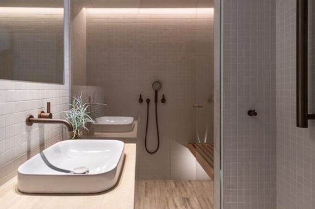 A refined contemporary bathroom featuring a large white vessel basin on a pale stone vanity, warm brass-toned tapware, pale small-format tiled walls, timber-look flooring, a walk-in shower with handheld fixtures, and a built-in timber bench. Soft concealed lighting and warm neutral finishes create a calm, spa-like atmosphere.