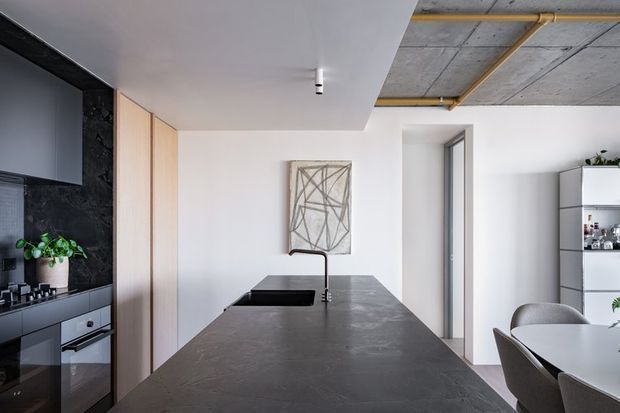 A contemporary open-plan kitchen viewed from the island, featuring a large dark stone island benchtop with integrated sink and black tap, matte dark cabinetry, pale timber pantry or storage doors, and a dark stone splashback. The adjacent dining area includes a round table and upholstered chairs, while exposed concrete ceiling panels and yellow service piping add an industrial character. White walls, minimalist shelving, and abstract wall art complete the space.