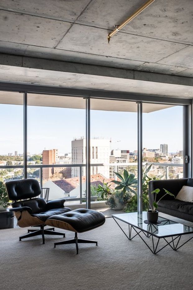 A modern high-rise living room with expansive floor-to-ceiling glazing and urban views. The space features a raw exposed concrete ceiling and beam, dark metal window frames, a black leather lounge chair with ottoman, a dark sofa, a geometric black coffee table with glass top, and indoor plants. The room has a refined mid-century-inspired furnishing scheme with strong industrial architectural elements and abundant natural light.