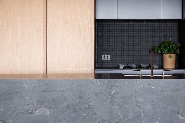 Close-up view of a contemporary kitchen featuring pale timber cabinetry, dark square-tile splashback, dark upper cabinets, a grey stone island or benchtop, and a brass-toned tap with a potted plant.