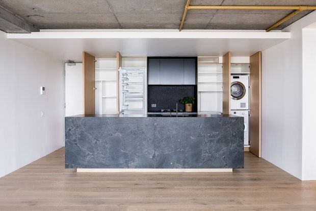 Open-plan kitchen with a large dark grey stone island and integrated cabinetry. The compact galley-style kitchen includes a black splashback, light timber shelving and panels, concealed appliances including a washing machine, and pale timber flooring. The space has white walls and an exposed concrete ceiling with visible yellow service pipework, creating a refined industrial feel.