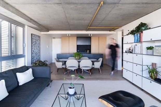 Open-plan contemporary living and dining area with a dark sofa, sculptural coffee table, dining table and chairs, extensive built-in timber and white cabinetry, and a black kitchen or media joinery wall. The space has exposed raw concrete ceilings with visible yellow conduit, large louvred windows, light timber flooring, and abundant indoor plants.
