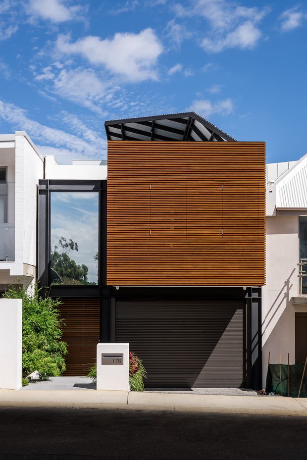 Street-facing exterior view of a narrow contemporary multi-storey residence. The facade combines a warm horizontal timber screen, dark metal framing, a large reflective window, and a dark roller garage door. A pitched, exposed roof structure projects above the timber-clad upper level, with neighbouring attached homes visible on either side.