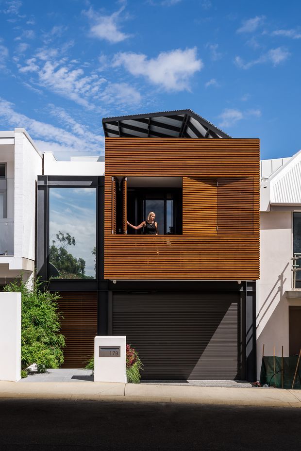 Street-facing view of a narrow multi-storey urban townhouse with a warm timber slatted upper facade, black-framed glazing, a dark garage door, and a steeply pitched exposed roof structure. The contemporary facade sits tightly between neighbouring homes under a bright blue sky.
