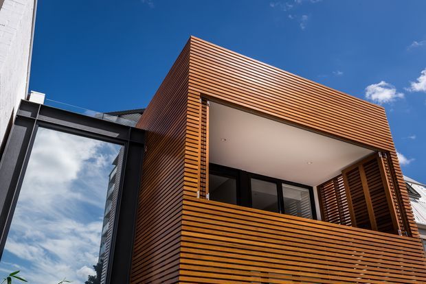 Upward-looking exterior view of a contemporary multi-storey building extension or facade. The main volume is clad in closely spaced horizontal timber slats, forming a projecting upper-level balcony or recessed outdoor space with dark-framed glazing and timber privacy screening. A contrasting dark-framed glazed section is visible at left, alongside part of a neighbouring or adjoining white facade. The scene is brightly lit under a clear blue sky.