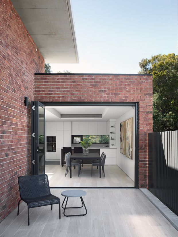 A contemporary brick courtyard or terrace viewed from outdoors, looking through a wide open glazed doorway into a bright open-plan kitchen and dining area. The exterior has red brick walls, a pale concrete overhang, dark metal fencing, and light grey tiled paving. Inside are white full-height kitchen cabinets, a dark dining table with chairs, a large artwork, and a skylight or recessed ceiling detail. A black outdoor lounge chair and small round side table occupy the foreground.