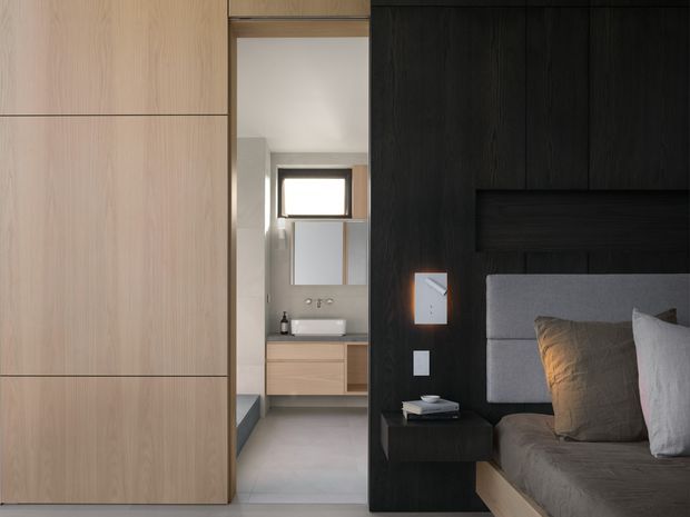 A refined modern bedroom viewed beside an open bathroom doorway. The bedroom features a dark vertical timber feature wall with an integrated recessed headboard and floating bedside table, a soft grey upholstered bedhead, neutral bedding, and warm wood flooring or joinery. The adjacent compact bathroom has pale cabinetry, a white basin, grey floor and wall surfaces, and a small horizontal window. Full-height pale timber wardrobe or sliding wall panels frame the opening.