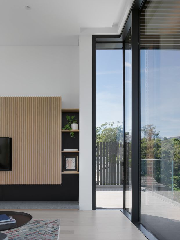 Bright contemporary living room with a minimalist built-in entertainment wall featuring vertical light-wood slatted panelling and black open shelving. Full-height black-framed glazing and an open glass door lead to a balcony or terrace, bringing in expansive daylight and views of trees and neighbouring rooftops. Pale timber flooring, white walls and ceiling, and restrained furnishings create a clean, airy interior.