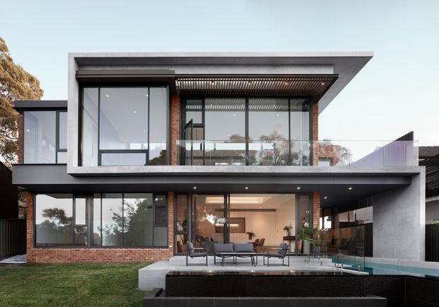 Exterior view of a large two-storey contemporary residence with a flat roof, extensive floor-to-ceiling glazing, dark projecting roof slabs, exposed red brick walls, concrete surfaces, and a covered outdoor entertaining terrace beside a swimming pool. The open-plan living area is visible through the glazing, with a balcony and upper-level screened shading structure.