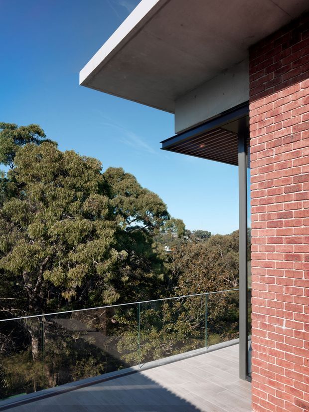 Exterior architectural photograph of a contemporary upper-level balcony or terrace overlooking mature trees. The composition highlights a projecting flat concrete roof slab, red brick facade, dark-framed glazing or doors, a timber-lined projecting awning, and a transparent glass balustrade over large-format pale paving.