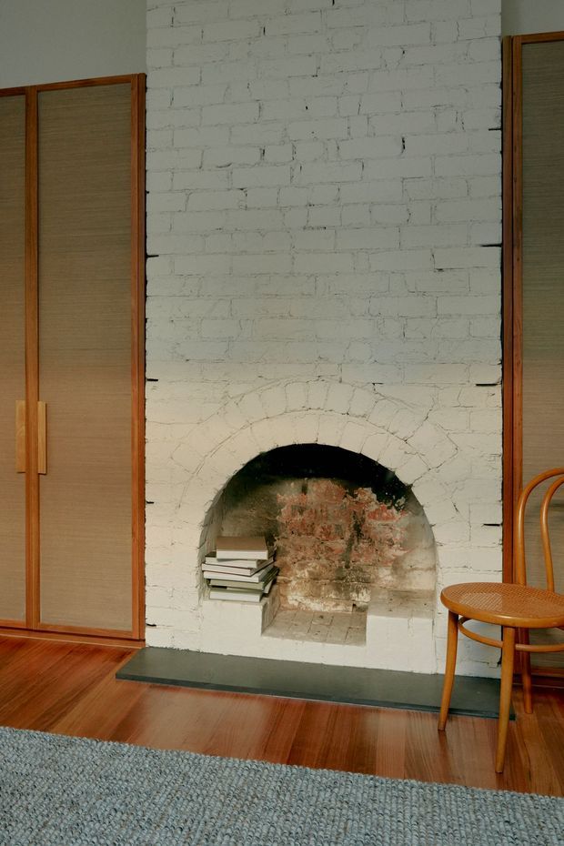 Interior detail of a warm, characterful room featuring a white-painted exposed brick wall with a shallow arched fireplace opening, weathered brickwork inside the firebox, and a dark stone hearth. Tall timber-framed built-in cupboards with woven or textured panel fronts flank the fireplace. A bentwood-style timber chair, stacked books, polished timber flooring, and a textured blue-grey rug complete the scene.