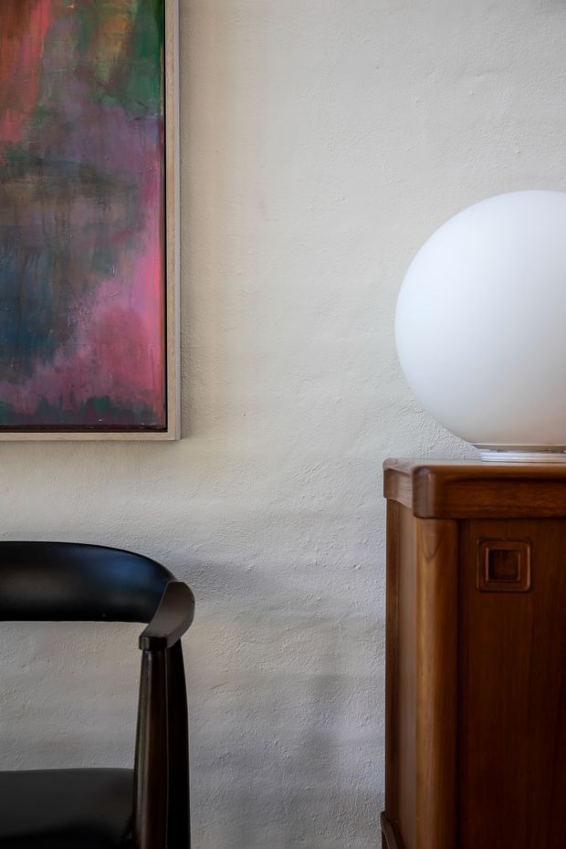 A close-up interior vignette showing a textured off-white plaster wall, a framed abstract artwork, a black sculptural chair, and a dark timber cabinet topped with a large white spherical lamp. The composition has a refined, gallery-like character with warm wood and dramatic black accents.