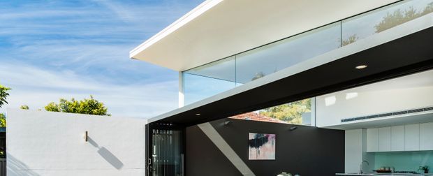 A contemporary flat-roof residence viewed from the exterior, featuring a strong cantilevered white upper volume, expansive frameless glazing, and a contrasting black structural or entry canopy. A partially visible minimalist kitchen can be seen through the glazing, while a white rendered boundary wall and clear blue sky reinforce the clean modern composition.