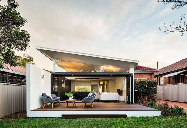 A contemporary single-storey rear extension opening onto a landscaped backyard. The pavilion-like addition has a dramatic sloping roof, extensive black-framed glazing and fully openable glass doors, with a timber deck furnished as an outdoor sitting area. A bright, modern living room with a sofa, armchairs, coffee table and built-in cabinetry is visible inside. Adjacent traditional brick homes and tiled roofs frame the extension.