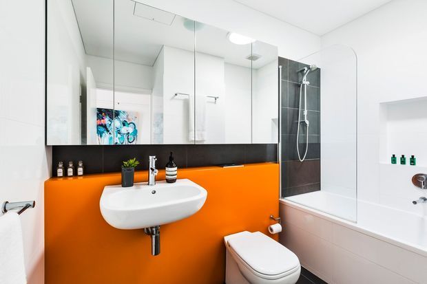 A bright contemporary bathroom featuring a wall-mounted white basin and toilet, a vivid orange painted feature wall beneath large mirrored cabinets, black tiled shower detailing, and a white bathtub with glass shower screen. The space has clean lines, built-in shelving, chrome fixtures, and strong black-and-white contrast with an orange accent.