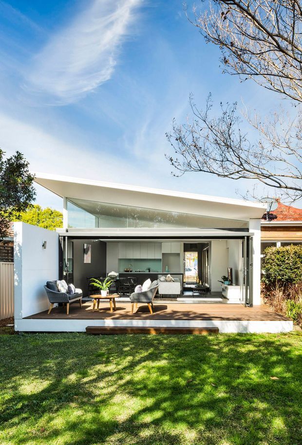 A bright contemporary rear extension opening onto a landscaped lawn. The single-storey structure has a crisp white flat roof with deep overhangs, extensive floor-to-ceiling glazing, large retractable glass doors, and a raised timber deck. Through the open facade is a combined indoor-outdoor living area with lounge seating, a small table, and a compact kitchen or bar area. Mature planting and neighbouring residential rooflines frame the suburban setting.
