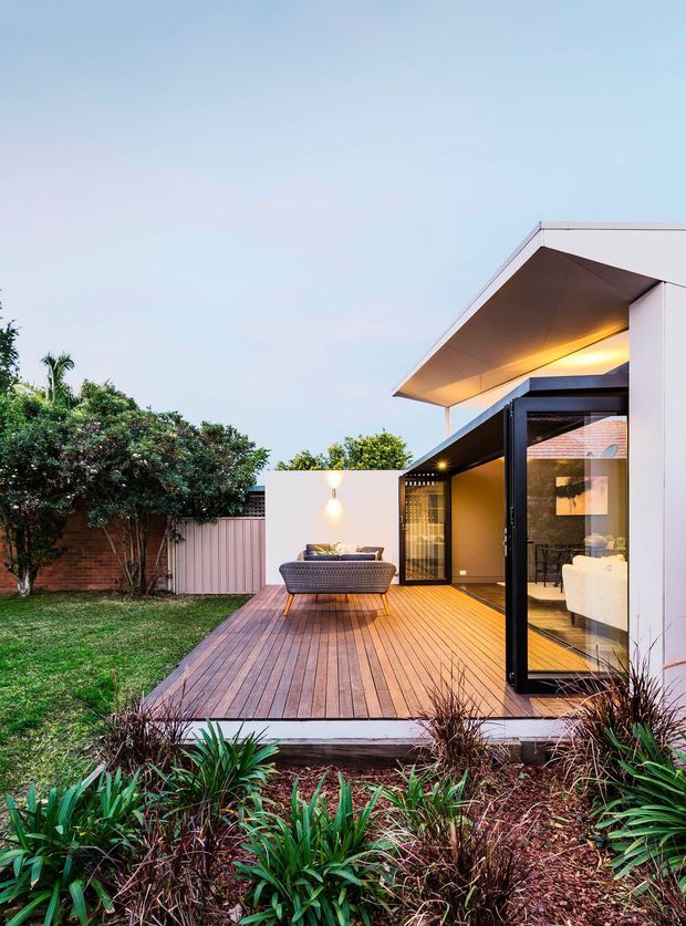 Exterior view of a contemporary single-storey home extension with a broad sloping roof canopy, white rendered walls and dark-framed glazed doors opening onto a raised timber deck. The deck includes outdoor seating and overlooks a landscaped garden, while the interior living area is visible through the glazing.