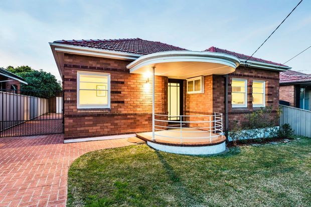 Exterior view of a compact single-storey red brick bungalow with a dark tiled hipped roof, curved covered front porch, metal handrails and a small semicircular entry platform. A red brick path leads past a gated side area, with a modest lawn and neighbouring residential structures visible.