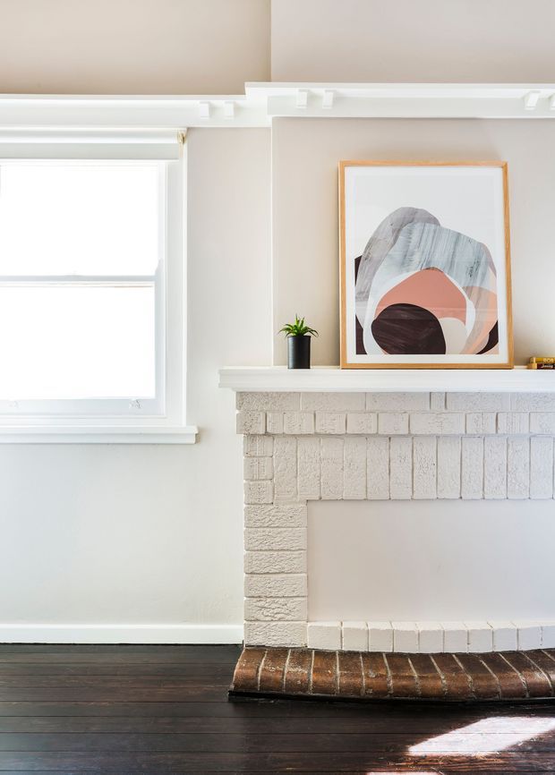 Bright close-up interior view of a traditional painted brick fireplace with a simple mantel, white-trimmed window, dark timber flooring, and neutral walls. A framed abstract artwork and small potted plant sit on the mantel, creating a clean, warm, understated living space.