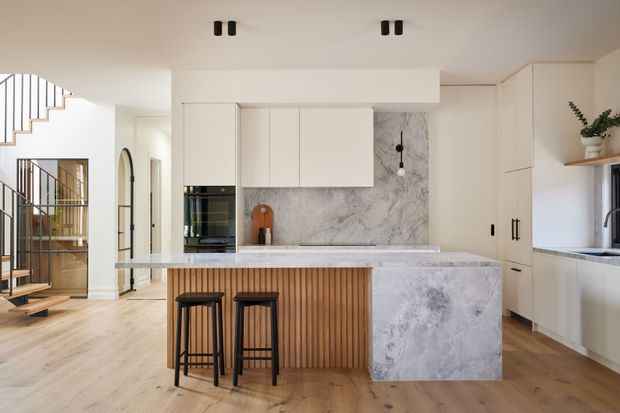 Bright contemporary kitchen with a large marble-look island, integrated white cabinetry, a veined stone splashback, timber-slatted island base, and two black bar stools. Light timber flooring and a visible staircase and hallway connect the kitchen to the rest of the home.