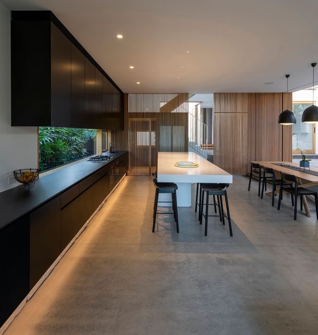 Open-plan contemporary kitchen and dining area with dark full-height cabinetry along the left wall, a large white island with bar seating, and a dining table beneath black pendant lights. Vertical timber screening and slatted wall elements divide the space, while a planted courtyard is visible through the kitchen glazing. The room has a polished, minimalist fit-out with a concrete-look floor and warm integrated lighting.