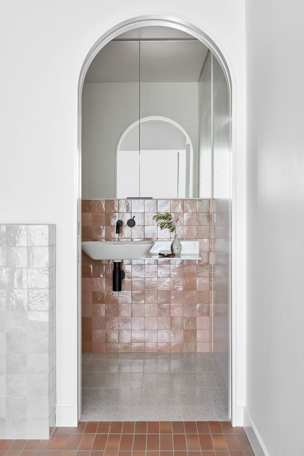 Bright compact bathroom viewed through a tall arched doorway. The room features a wall-mounted white basin with matte black tapware, a large arched mirror, glossy blush-pink square wall tiles, and a grey terrazzo-style floor. White curved architectural trim frames the opening, while terracotta-toned floor tiles continue outside the bathroom. A small vase with greenery sits on the basin.
