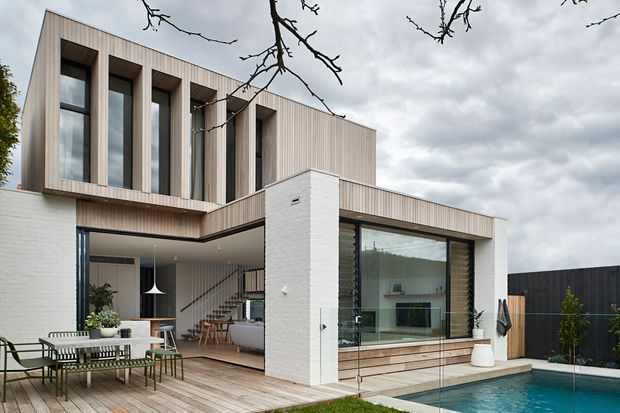 Contemporary two-storey residence with a pale vertically clad upper level, white masonry walls, extensive glazing, and a timber-lined outdoor entertaining terrace. The rear elevation opens onto a deck with dining furniture and a swimming pool, while the interior living and dining area is visible through the glazing. The composition has strong horizontal and vertical geometric forms and a restrained natural palette under an overcast sky.