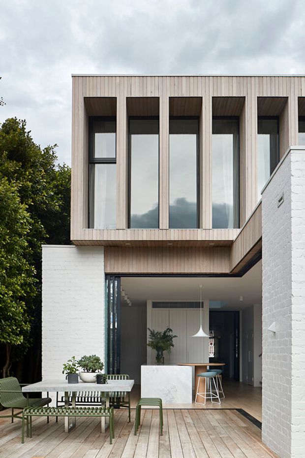 A contemporary two-storey residential facade opening onto a timber deck and outdoor dining area. The upper level is clad in vertical light timber boards and features tall, deeply recessed dark-framed windows. White-painted brick walls frame a partially covered ground-floor patio, with a compact kitchen visible inside. The scene includes outdoor furniture, built-in-looking planting, and a covered indoor-outdoor connection.