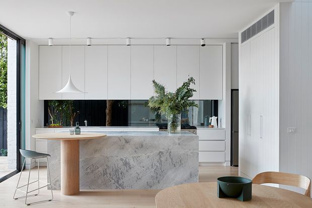 Bright, open-plan contemporary kitchen and dining area with a large marble-look island, white full-height cabinetry, integrated appliances, timber flooring, and expansive glazing. A sculptural white pendant hangs above the island, while a timber dining table and chairs occupy the foreground. The space has a clean, refined, minimalist feel with abundant natural light.