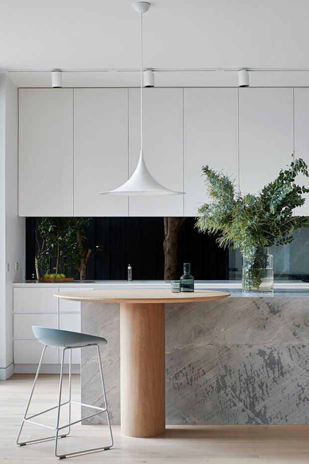 A refined contemporary kitchen centred on a rounded timber island with a cylindrical pedestal base and pale stone benchtop. The island front has a striking grey textured stone or plaster finish, while the surrounding cabinetry is handleless and white. A sculptural white pendant hangs above the island, complemented by recessed track spotlights, a pale timber floor, a pale blue bar stool, and large leafy branches in glass vessels. Dark vertical panelling and indoor planting add contrast behind the kitchen.