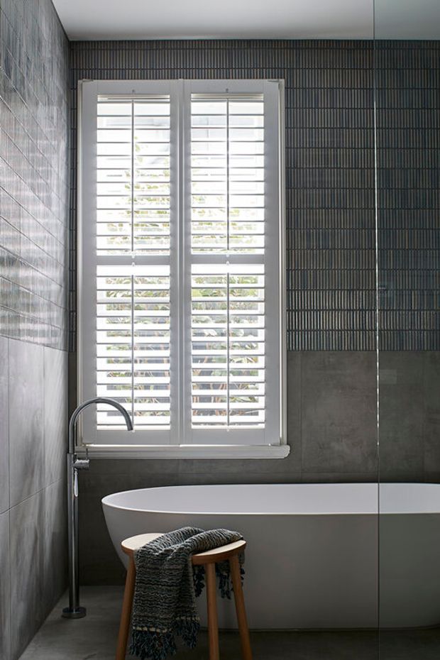 A compact, refined bathroom featuring a freestanding white bathtub beneath a large white plantation-shutter window. Dark grey textured wall tiles and concrete-look surfaces create a moody contemporary backdrop, with a slim freestanding bath filler and a small wooden stool draped with a dark woven throw. A frameless glass shower screen is visible at the right edge.