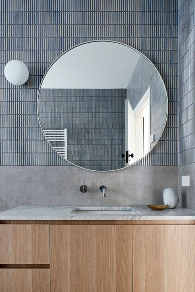 Close-up view of a contemporary bathroom vanity. The wall is clad in narrow vertically ribbed grey tiles, with a large round mirror and spherical wall light above a wall-mounted basin tap. A pale stone benchtop sits over light timber-grain vanity cabinetry, with a small decorative bowl and vessel at the side. The mirror reflects a shower or adjacent tiled area and a doorway.