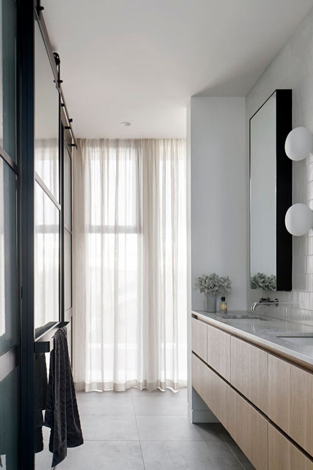 A bright, narrow contemporary bathroom with a long floating timber vanity, pale stone benchtop, integrated basin and wall-mounted tapware. A large black-framed mirror with spherical wall lights sits above the vanity, while a black-framed glass shower enclosure and dark towel occupy the left side. Pale large-format floor tiles, vertical light wall tiles, and sheer curtains create a clean, softly lit interior.