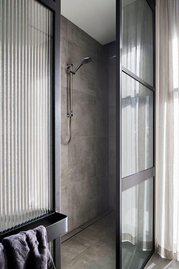 A narrow, contemporary walk-in shower area viewed from a bathroom. Large grey concrete-look tiles cover the shower walls and floor, with a minimalist chrome handheld shower fitting. Black-framed ribbed glass partitions and a black-framed glass door create a strong geometric enclosure, while sheer curtains allow soft daylight into the space. A partial black vanity or basin and folded towel are visible in the foreground.