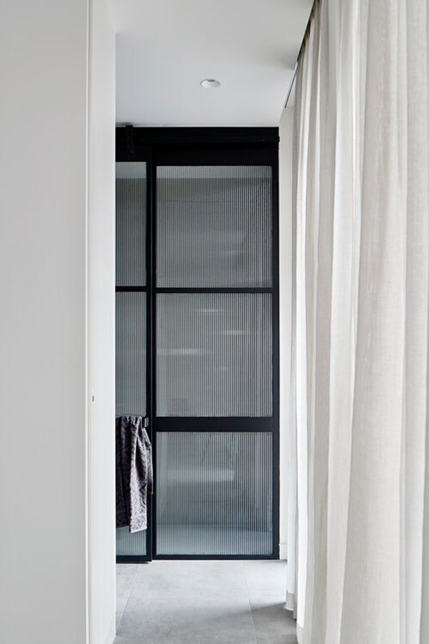 A narrow, minimalist bathroom viewed through a doorway, featuring a black-framed ribbed or reeded glass shower enclosure, pale tiled flooring, white walls and ceiling, and sheer white curtains along the right side. A dark towel hangs on the enclosure frame. The space has a restrained monochrome palette and clean contemporary detailing.