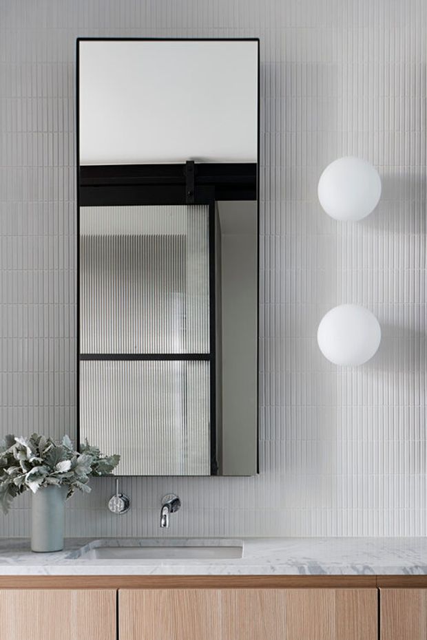 A refined contemporary bathroom vanity featuring vertically ribbed white wall tiles, a large black-framed mirror with a reflected fluted glass panel, two spherical wall lights, a marble-look benchtop with integrated basin, chrome wall-mounted tapware, and warm timber cabinetry below. A small vase of foliage adds a natural accent.