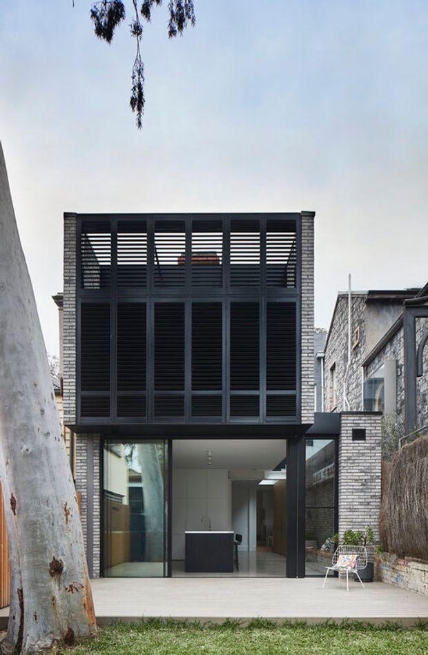 A narrow contemporary urban house extension viewed from a small rear garden or patio. The two-storey facade combines pale grey brickwork with a dark framed upper-level screen of horizontal louvres and full-height glazing below. The ground floor opens onto a light timber deck, revealing a minimal white kitchen or living area. Adjacent masonry walls, a mature tree, and dense neighbouring buildings create an enclosed urban setting.