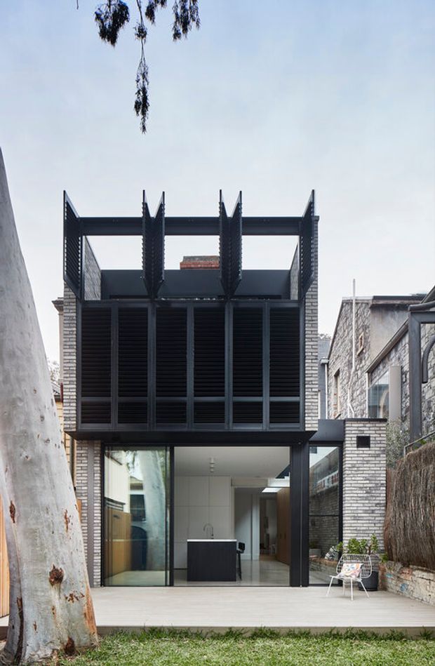 A narrow multi-storey urban house extension viewed from a small rear deck and garden. The facade combines pale brickwork with dark steel framing, extensive black louvres, and an exposed upper-level structural frame. Large ground-floor glazing opens to a minimalist kitchen and living area, while neighbouring masonry walls and mature trees frame the compact site.