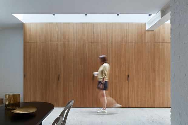 A minimalist interior corridor or open-plan living space with a full-height warm timber-clad wall of concealed storage or doors, a polished light concrete floor, and a large overhead skylight. A dark dining table and chairs occupy the foreground, while a person walks through the space. White painted masonry walls and restrained detailing create a calm, contemporary atmosphere.