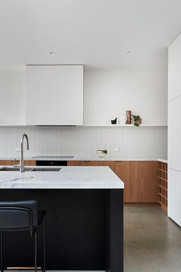 A bright, contemporary galley kitchen with a large black island topped in white veined stone, integrated sink and sculptural faucet, white tiled splashback, white brick wall, timber lower cabinetry, and a tall white appliance cabinet. A simple black bar chair sits at the island. Recessed ceiling lights and open shelving create a clean, restrained composition.