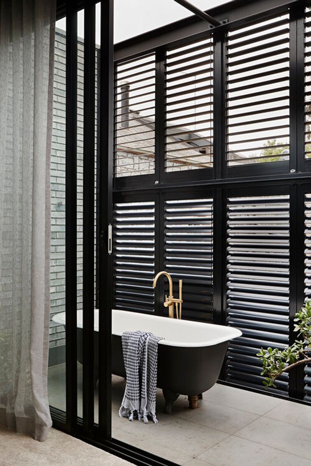 A contemporary bathroom featuring a freestanding black-and-white bathtub positioned beside a large black-framed glazed louvred screen. The space has pale grey floor tiles, a light grey brick wall, a sheer curtain, and a brushed brass wall-mounted bath tap. The louvres provide privacy and filtered daylight, with greenery visible at the edge of the frame.