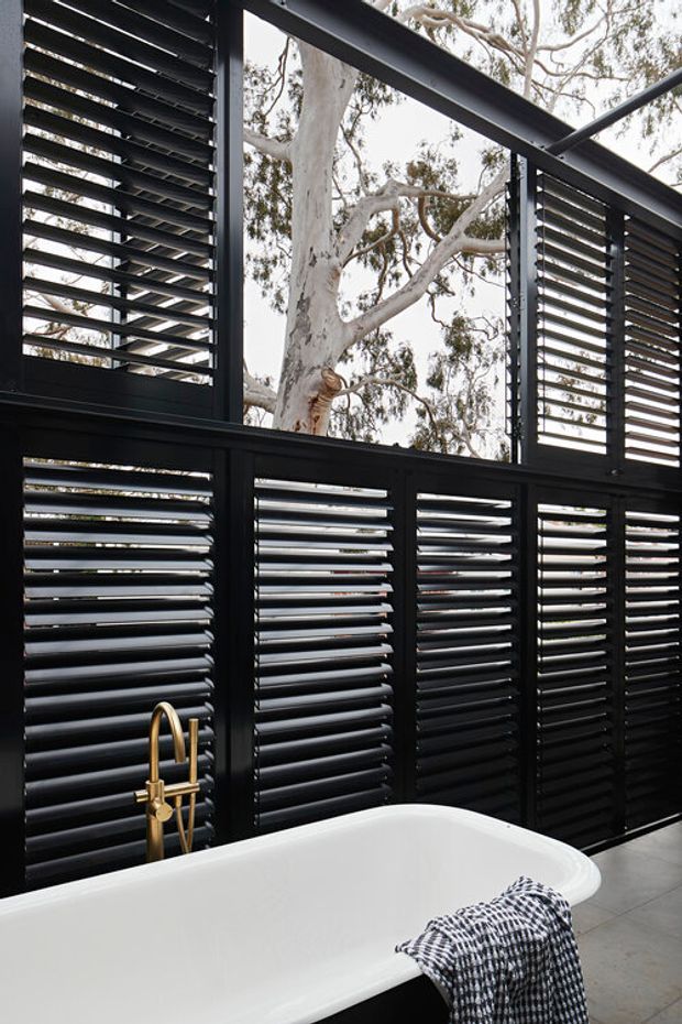 A contemporary bathroom featuring a white freestanding bathtub positioned against dramatic black louvred window and wall panels. Large openings provide views to eucalyptus trees, while a brushed brass/gold floor-mounted bath tap adds a warm accent. The space has a refined, high-contrast indoor-outdoor character with grey tiled flooring and abundant natural light.