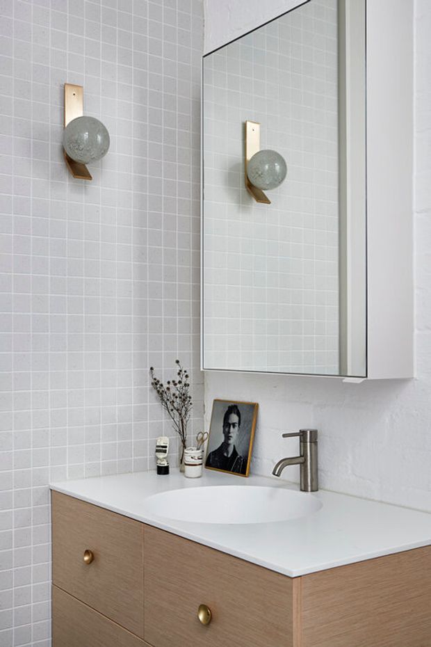 A compact, softly styled bathroom vanity with a white integrated basin, pale timber floating cabinetry, brushed metal tapware, and a large mirrored cabinet. The walls are clad in small pale grey square tiles, with a globe wall light on a brass-toned bracket and understated decorative accessories.